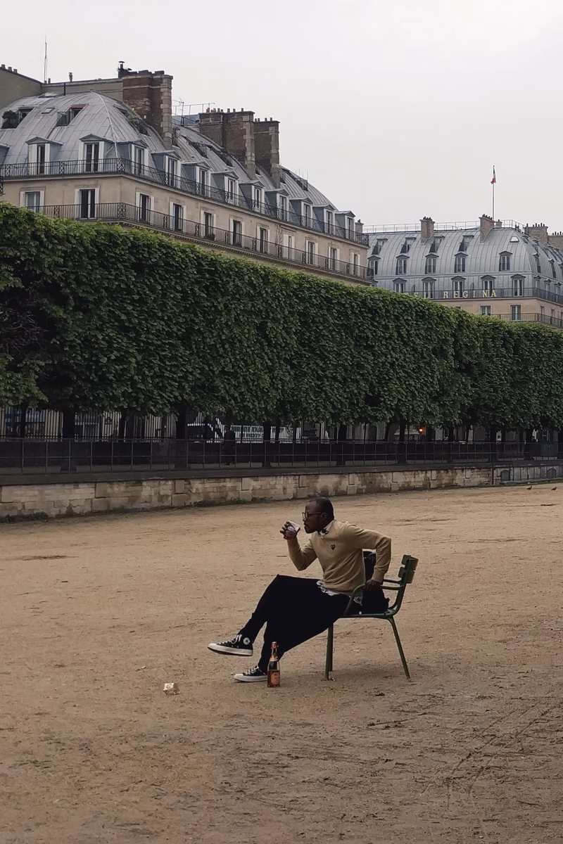 A man sits on a chair in a park, drinking from a bottle.