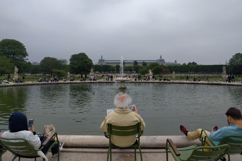 Several people sit by a pond with a fountain in the center, overlooking a large classical building under an overcast sky.