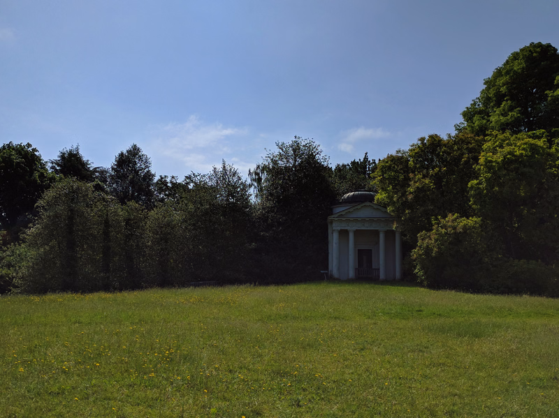 A small classical temple structure is visible in a grassy garden area surrounded by dense trees under a bright blue sky.