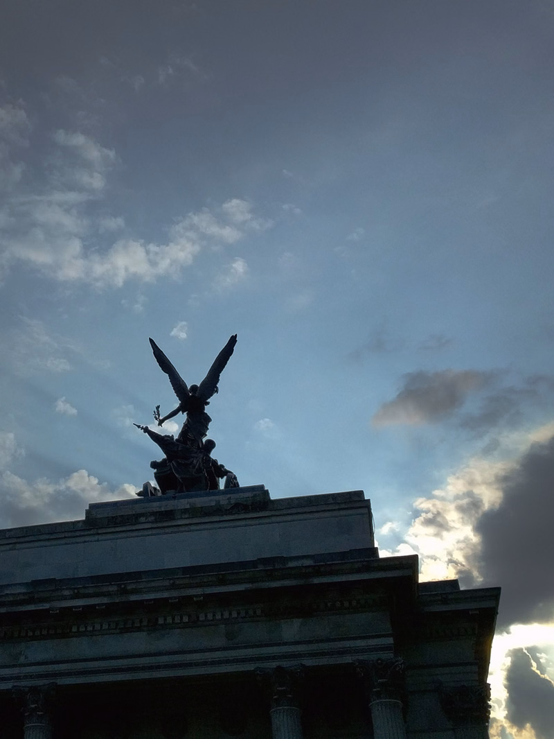 A dark silhouette of a statue atop a large building against a cloudy, overcast sky.