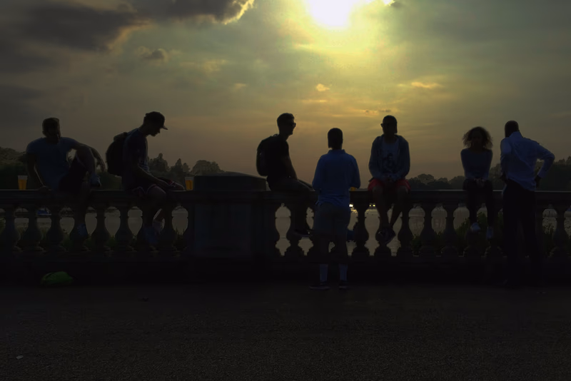 A group of people sitting and standing on a balcony overlooking a serene landscape with a setting sun.