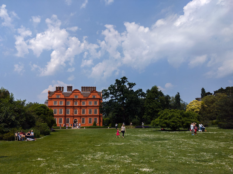 A large, red brick house is visible across a wide green lawn under a bright, cloudy sky.