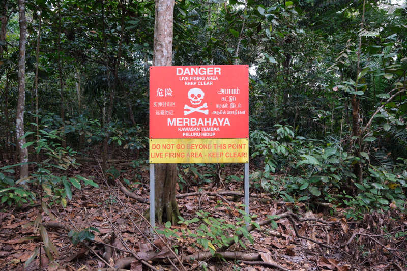 A warning sign in a forest reads "DANGER LIVE FIRING AREA KEEP CLEAR" in multiple languages, including English, Chinese, and Malay.
