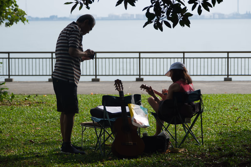 A man and a woman are playing a guitar and sitting on the grass near a body of water. They are surrounded by shopping bags and a fishing rod.