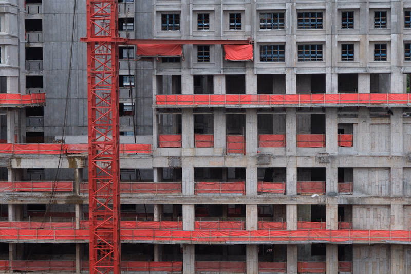 A tall, multi-story concrete building under construction features red scaffolding and exposed structural elements.