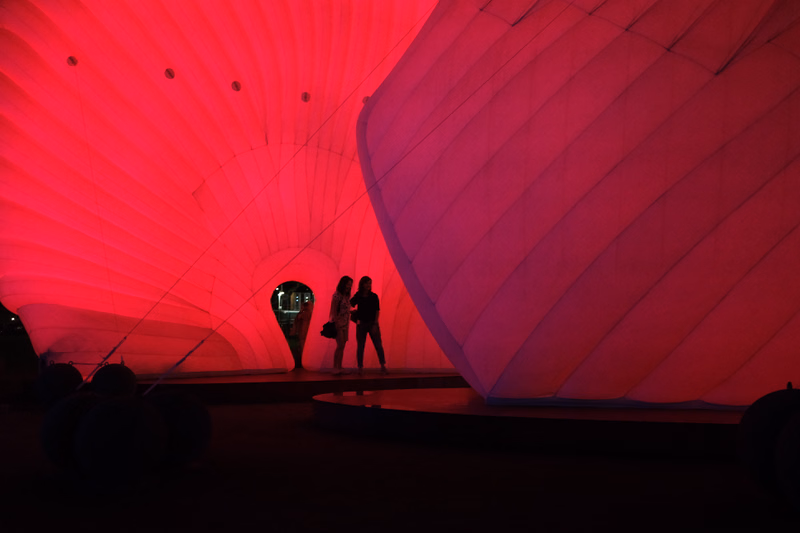 Three people stand on a stage inside a large, brightly lit red tent structure.