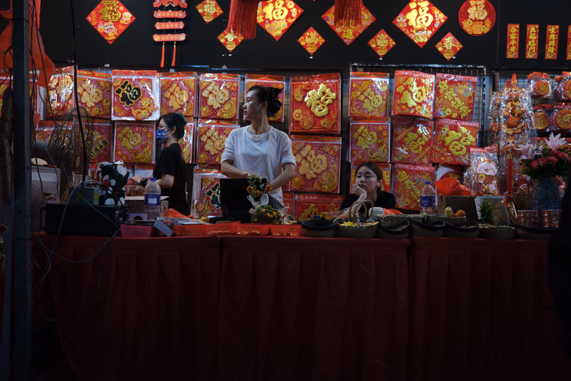 Several people stand behind a long red-draped market stall filled with brightly colored, ornate red and gold packages.