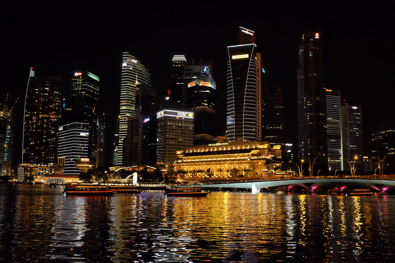 A nighttime cityscape featuring illuminated modern buildings reflected in the water of a river.