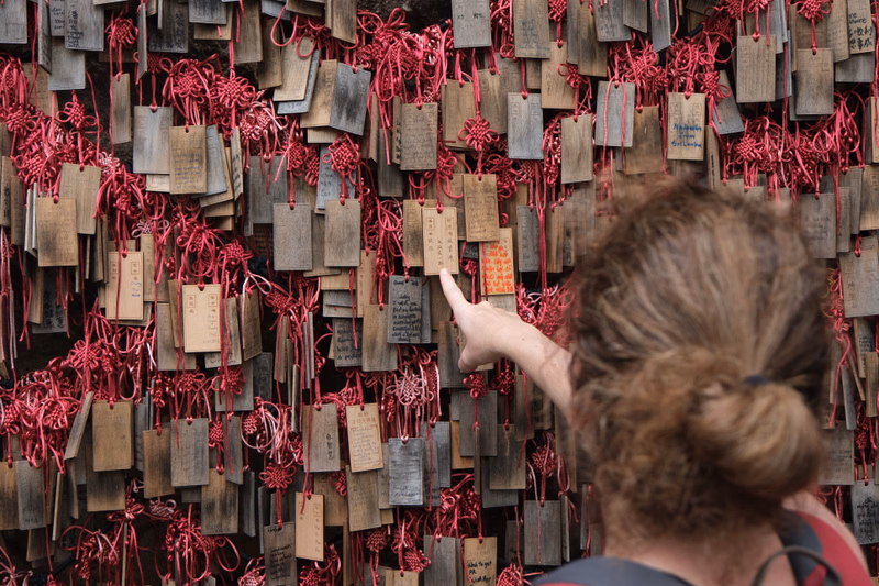 A person views a wall covered in numerous wooden plaques and bright red prayer ribbons.