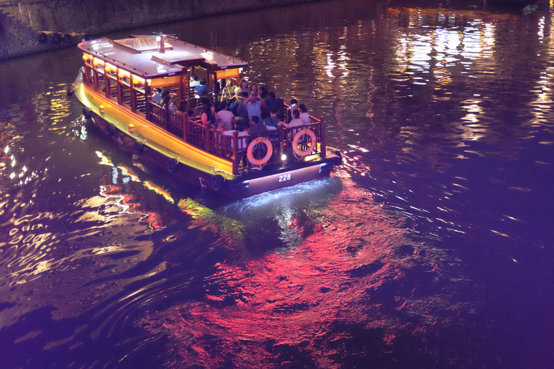 A yellow tour boat, numbered 228, travels on a dark river at dusk, with reflections visible on the water.
