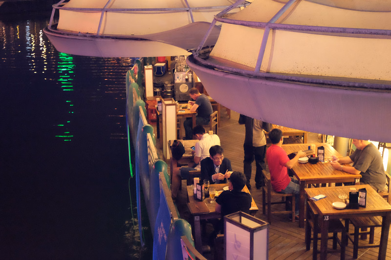 People are dining in an outdoor restaurant area covered by large white awnings next to a dark body of water.