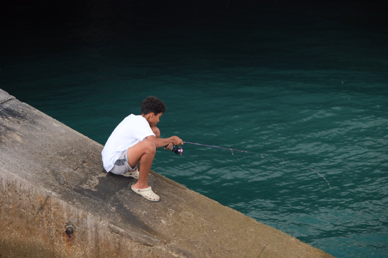 A young boy wearing a white shirt is fishing from a concrete pier into the dark green water.
