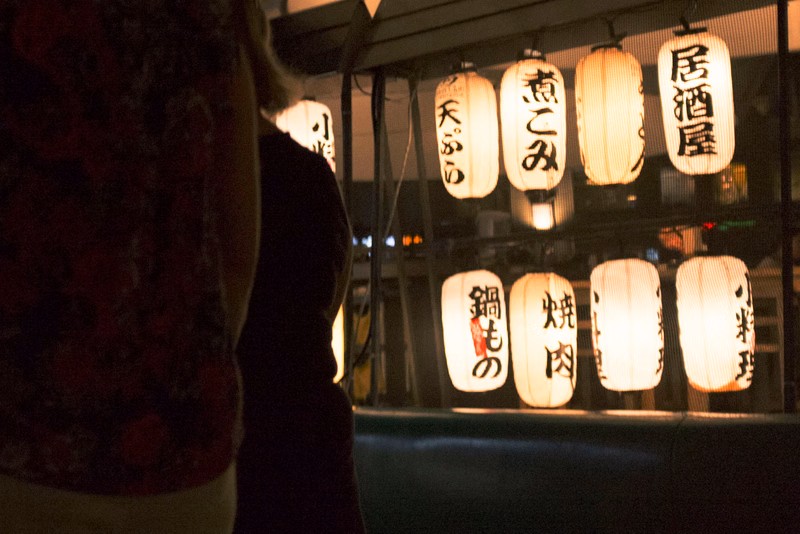 A row of illuminated Japanese lanterns hangs outside a shop, with visible text including "居酒屋" and "炙り肉".