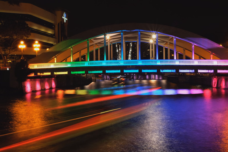 A modern arched bridge illuminated with colorful lights reflects vibrant streaks of light on the dark water at night.