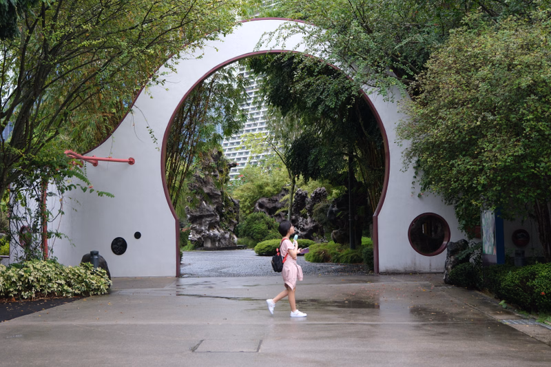 A woman in a pink dress walks through a large white archway into a lush garden area with a modern building visible behind the trees.