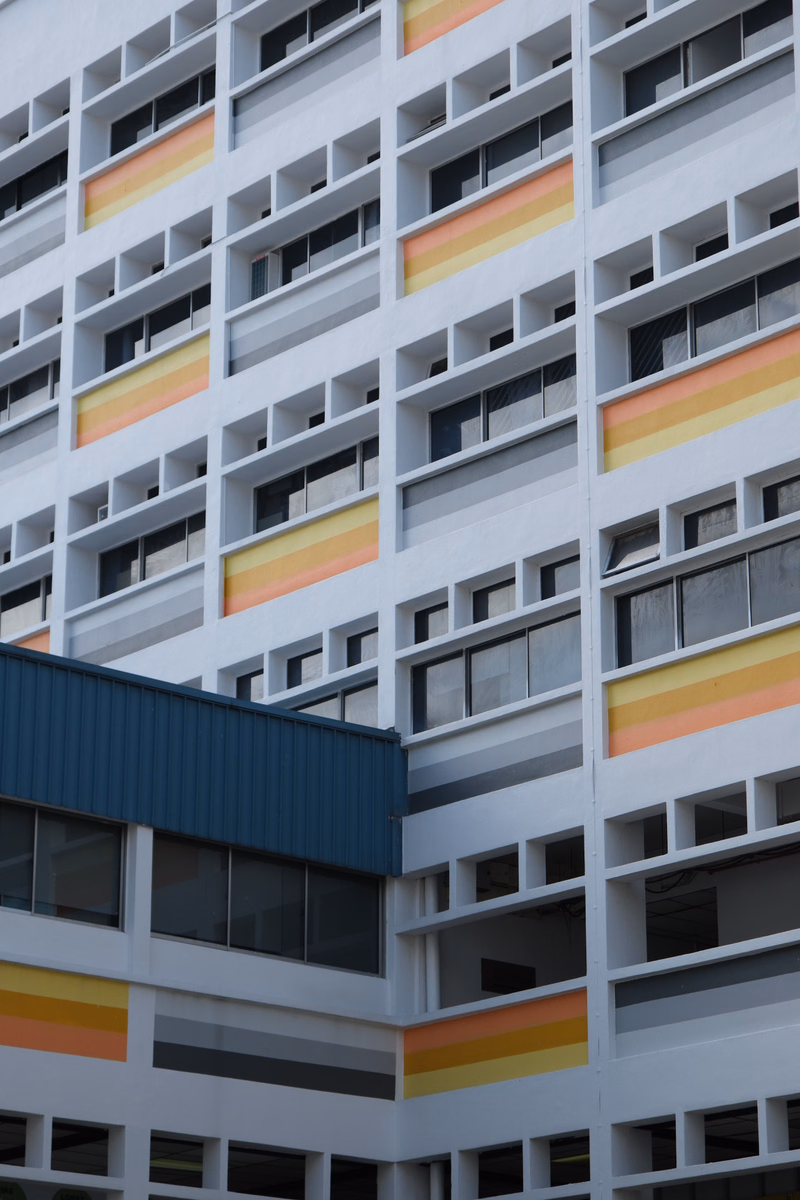 A modern, multi-story building facade features rows of windows interspersed with colorful horizontal panels in shades of yellow, orange, and pink.