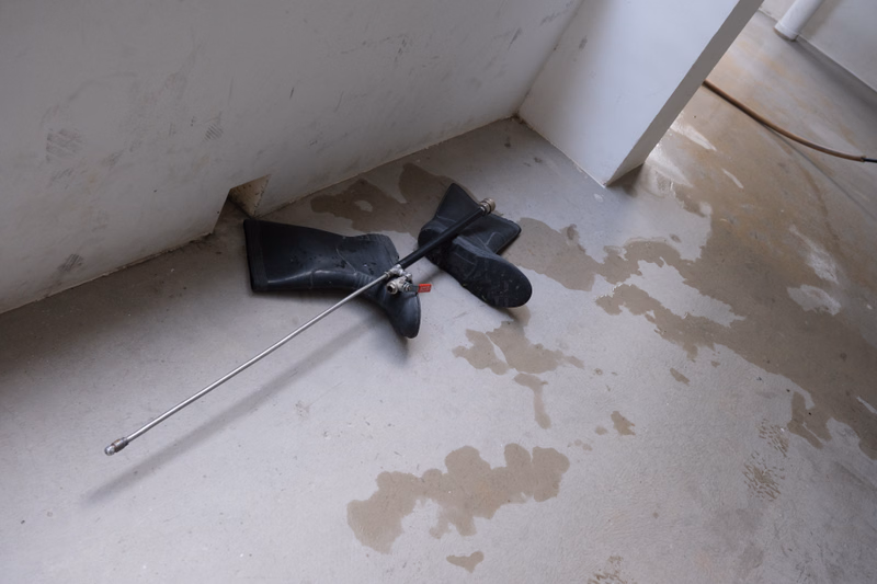 A metal pipe and several black plastic drain fittings are lying on a concrete floor near a white wall, with water pooling nearby.