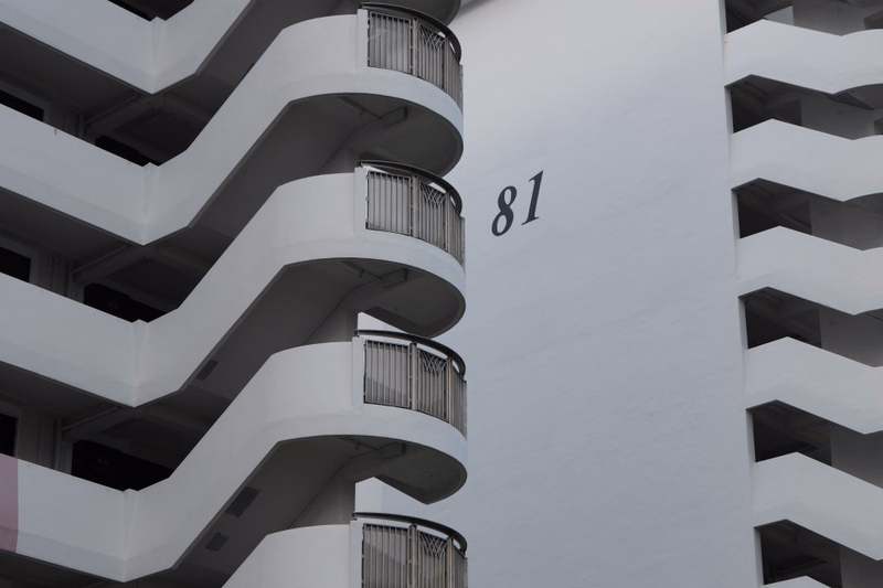 The white facade of an apartment building features curved balconies and the number "81" displayed on the wall.