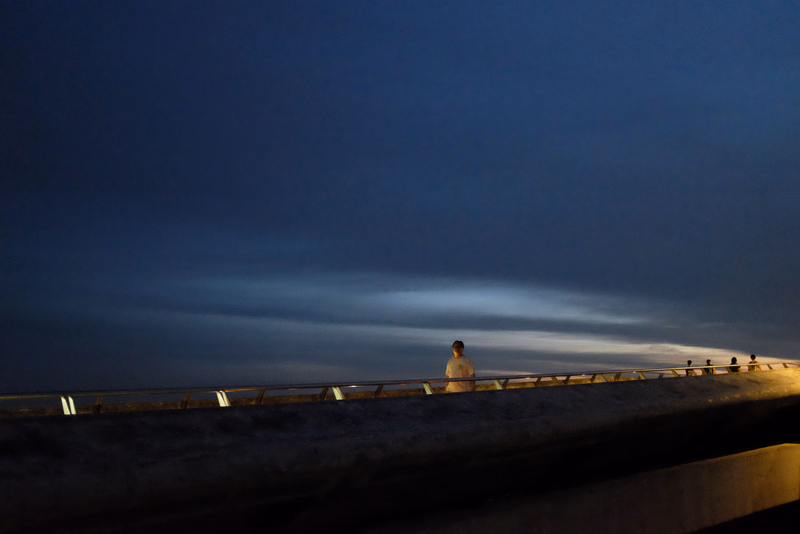 A lone person stands near a railing overlooking the ocean under a dark, twilight sky.