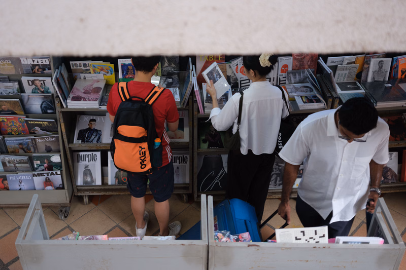 A man and a woman browse magazines in a shop display, with the man wearing an orange backpack.
