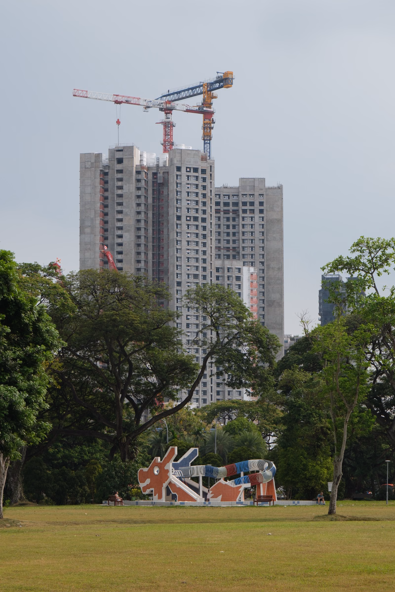A view of tall apartment buildings under construction with cranes, set behind a green park featuring a colorful abstract sculpture.