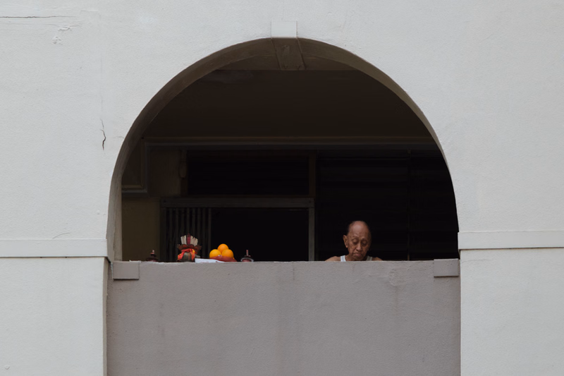 An elderly man looks out from a window opening beneath a large white archway.