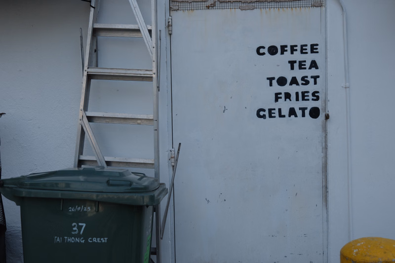 A white door has a menu painted on it that reads "COFFEE TEA TOAST FRIES GELATO", next to a green trash bin.