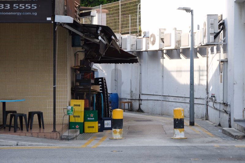 A row of external air conditioning units lines a white building exterior next to a dark awning and a yellow bollard.