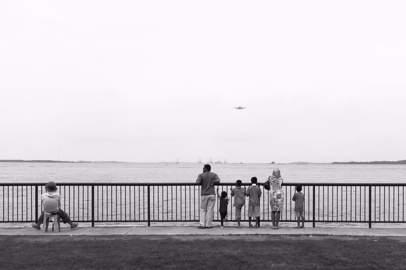 A family of six people standing by a fence overlooking a body of water, with a plane flying overhead.