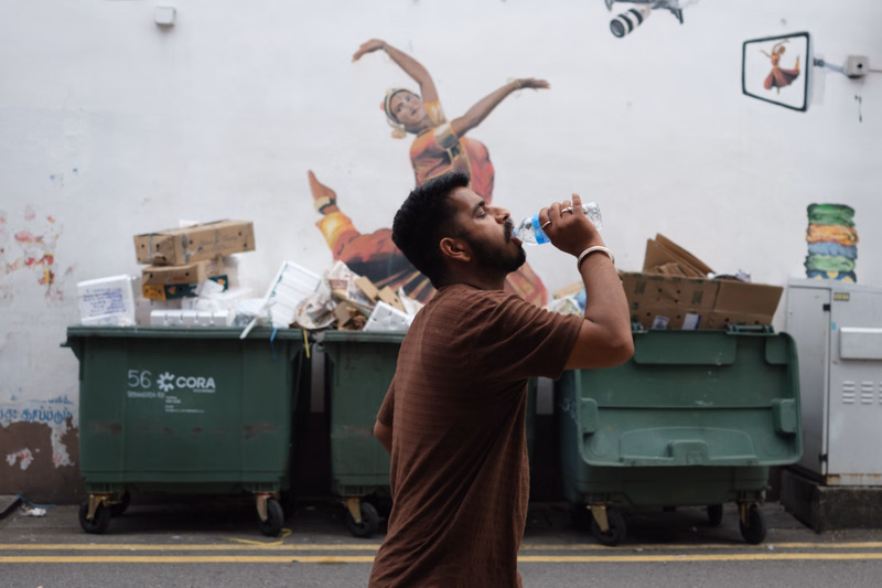 A man in a brown shirt drinks from a clear plastic bottle while standing in front of several green dumpsters with the text "56 CORA" visible.