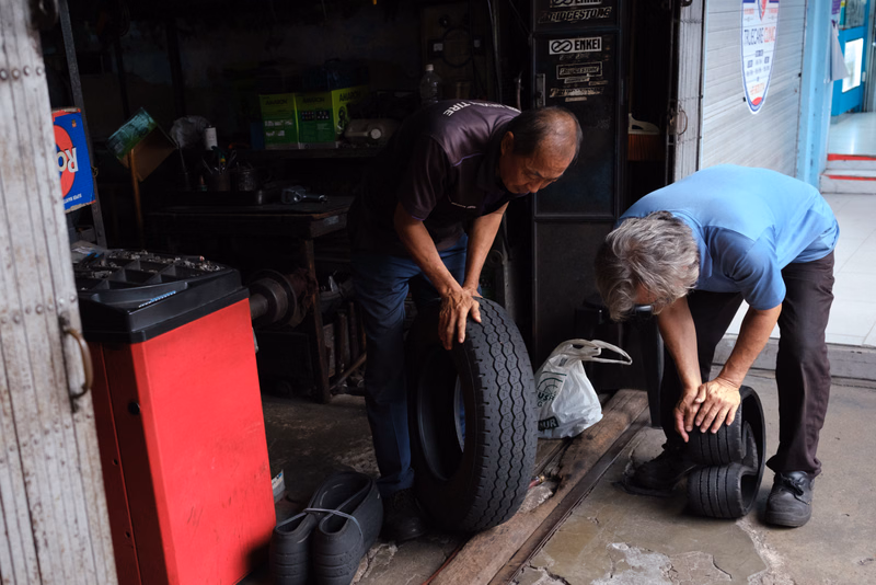 Two men in a workshop are working on a large tire, with tools and equipment visible in the background.