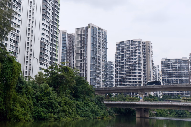 A row of modern apartment buildings stands behind a bridge over a body of water, surrounded by lush green trees under an overcast sky.