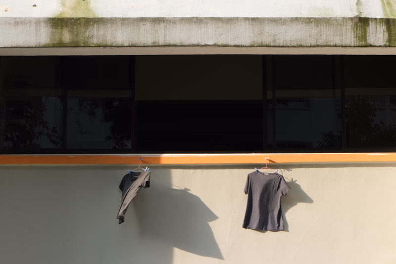 Two dark t-shirts are hanging on a clothesline against a light-colored exterior wall under a roof overhang.