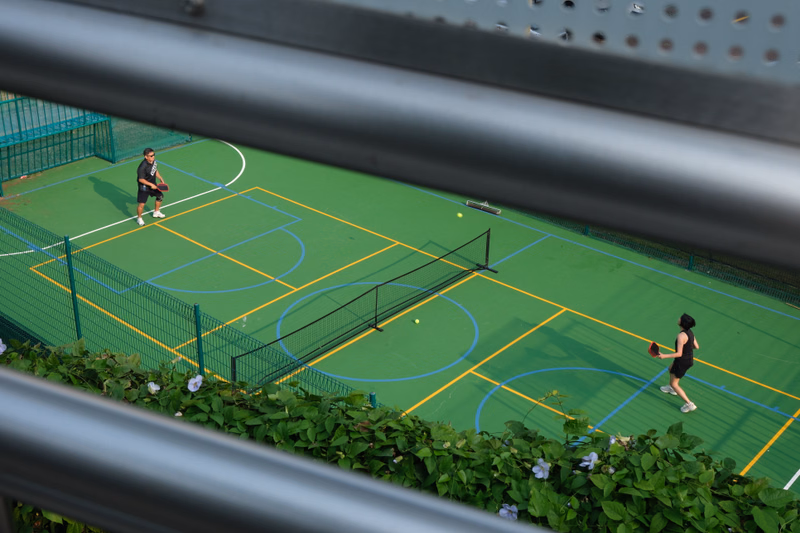 Two people are playing tennis on a green outdoor court, with a net dividing the playing area.