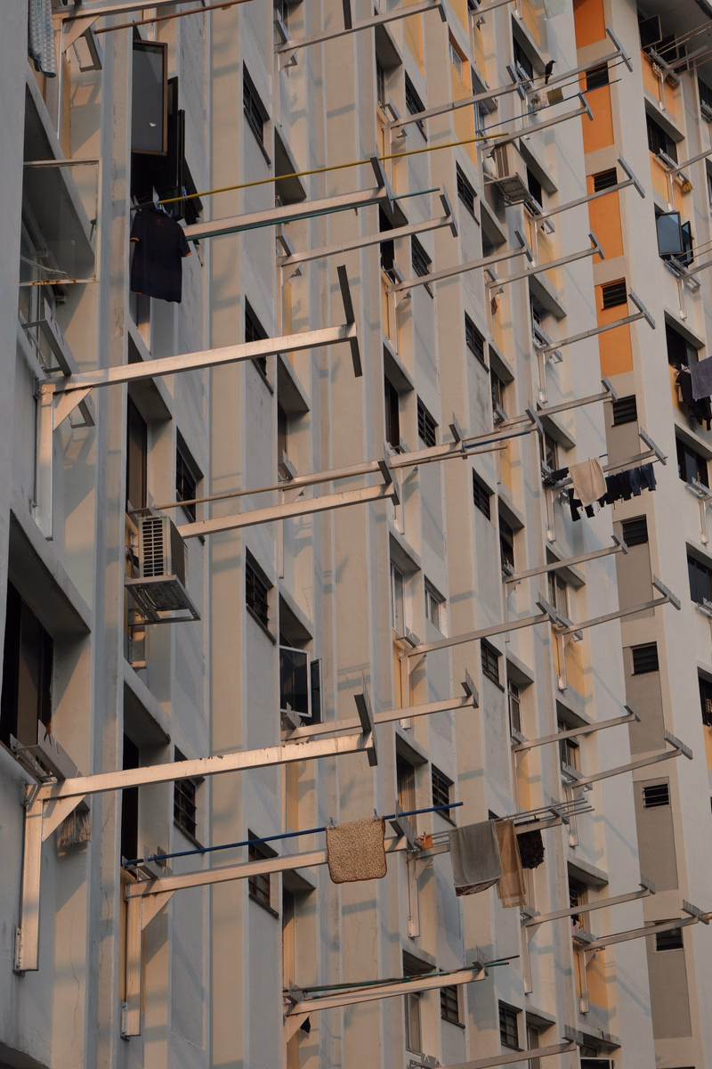 A tall, light-colored apartment building exterior features numerous balconies and many mounted air conditioning units.