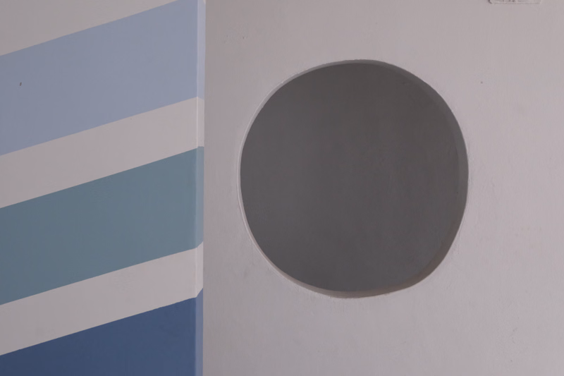 A close-up of a white wall featuring a circular window next to a section painted with horizontal stripes of light blue and white.