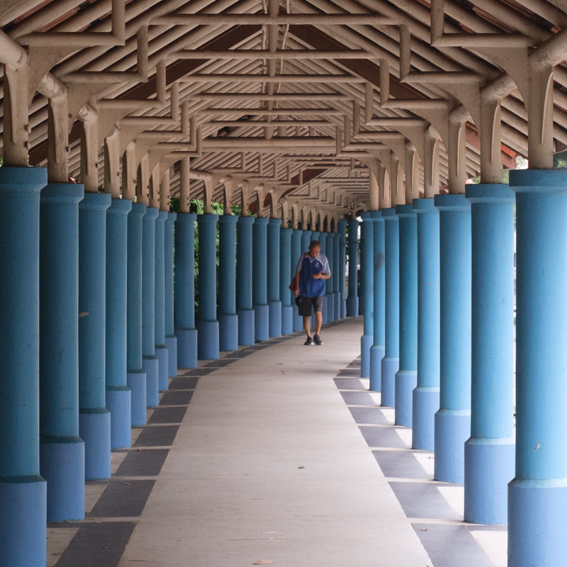 Two people walk down a long, covered walkway supported by numerous blue columns and a wooden roof structure.