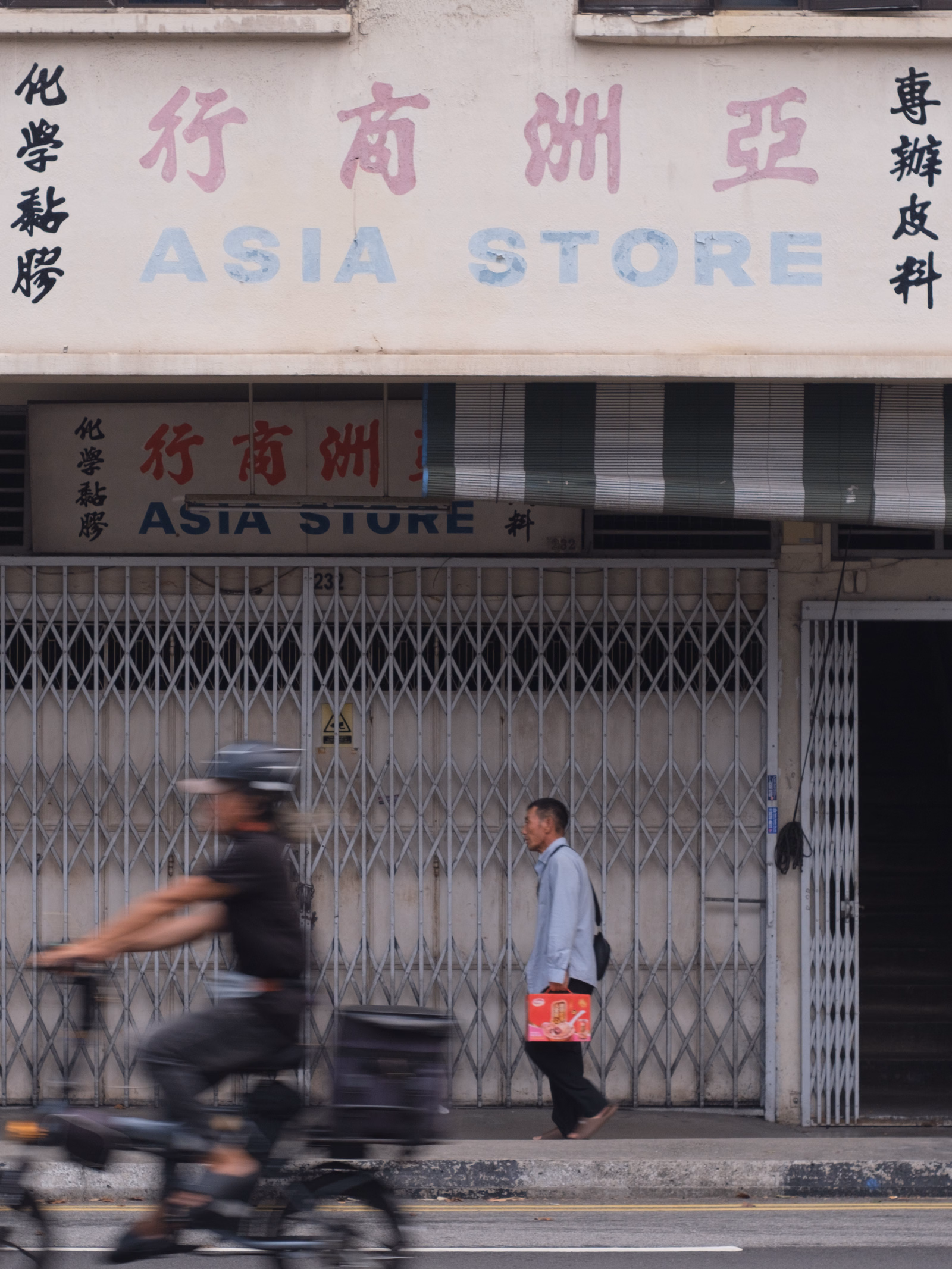 A storefront with Chinese and English text reading "仔南洲亞 ASIA STORE" is visible above a metal gate, with a person walking past and a person on a bicycle in the foreground.