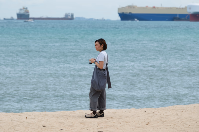 A woman stands on a beach, looking at the sea with a camera in her hand.