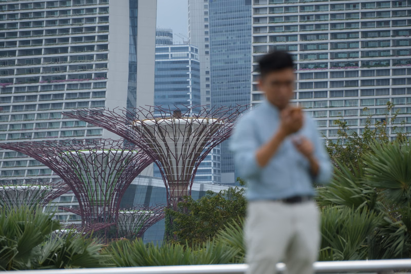 A man in a light blue shirt stands in the foreground against a backdrop of modern skyscrapers and a large, intricate metal art installation.