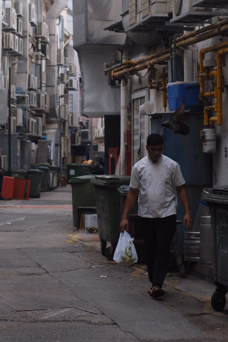A man in a white shirt walks down a narrow, urban street lined with apartment buildings and external air conditioning units.