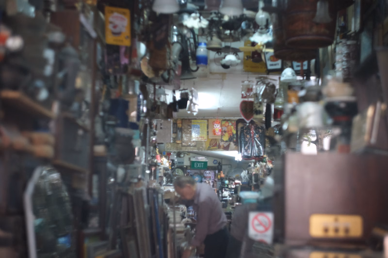 A cluttered indoor shop interior with many hanging goods and shelves, featuring an illuminated 'EXIT' sign in the distance.