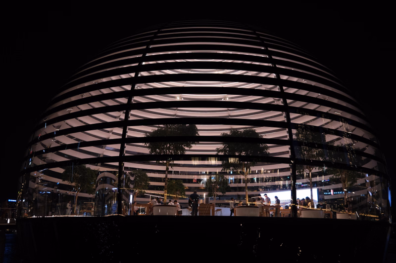 A large, illuminated spherical building at night features horizontal bands of light and visible trees inside.