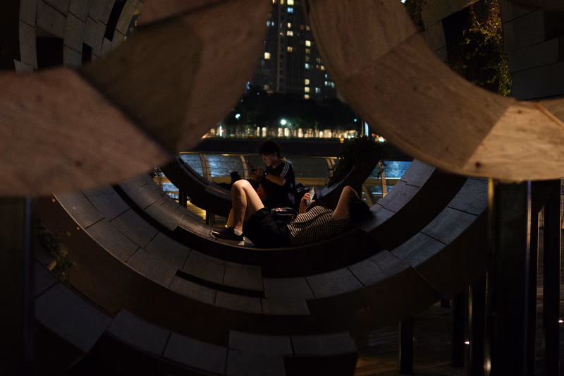Two people sitting on a circular structure near a body of water in a city at night.