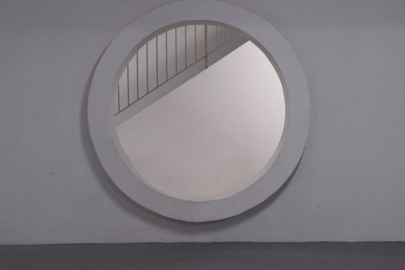 A large circular window with vertical bars is set into a plain white wall, looking into a bright interior space.