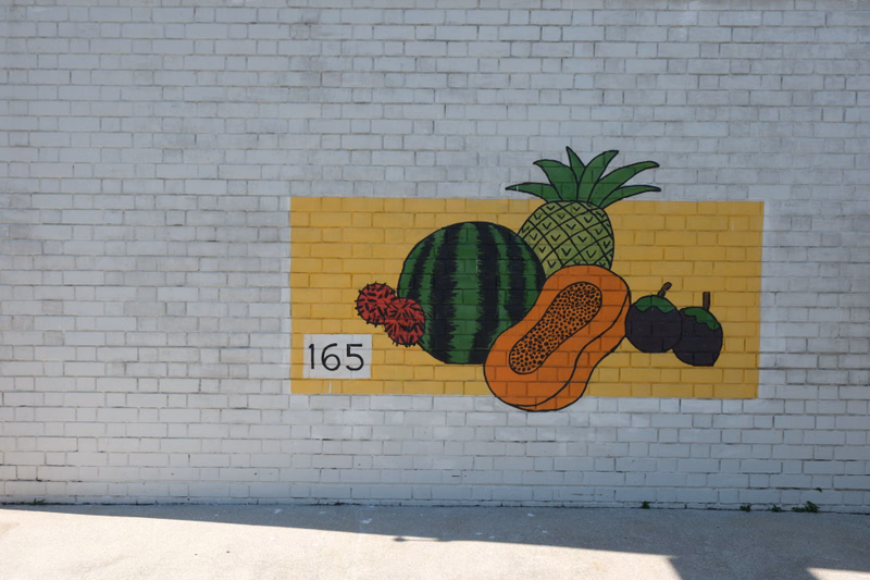 A colorful mural painted on a white brick wall featuring various fruits, including a watermelon, pineapple, pumpkin, and citrus.