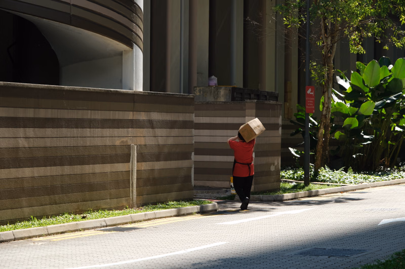 A woman wearing a straw hat walks along a paved walkway next to a modern building with tall columns and green foliage.