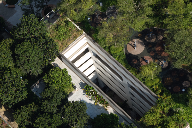 An aerial view of a modern, angular building surrounded by lush green trees and paved outdoor areas.