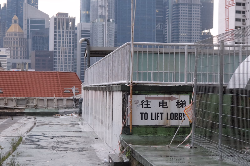A sign on a concrete structure reads "往电梯 TO LIFT LOBBY" next to a chain-link fence overlooking a waterfront area with tall buildings in the background.