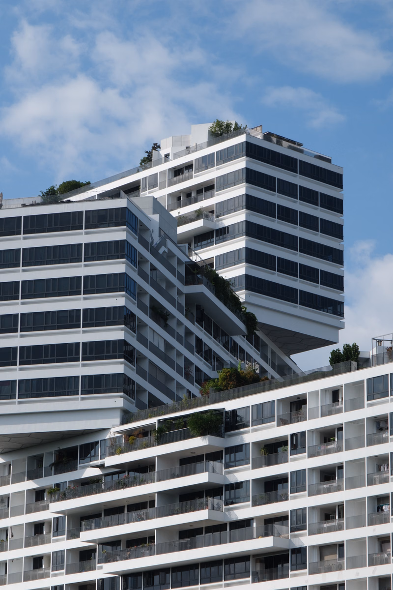 A modern, multi-story apartment building with numerous balconies is visible against a partly cloudy blue sky.